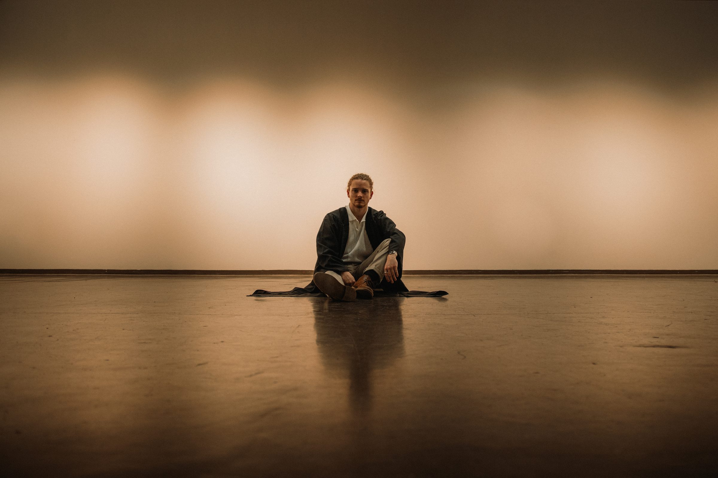 A man sitting on a reflective floor in front of a warmly lit empty wall