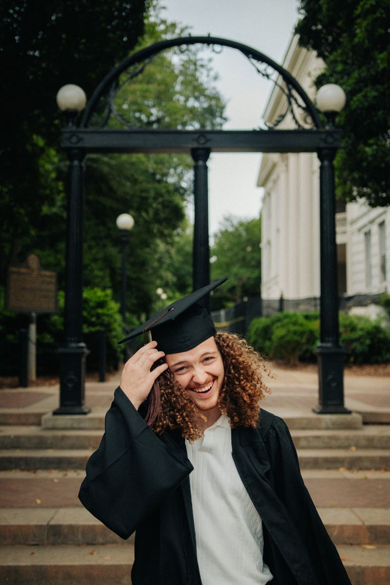 A graduate in cap and gown laughing beneath an iron arch on campus