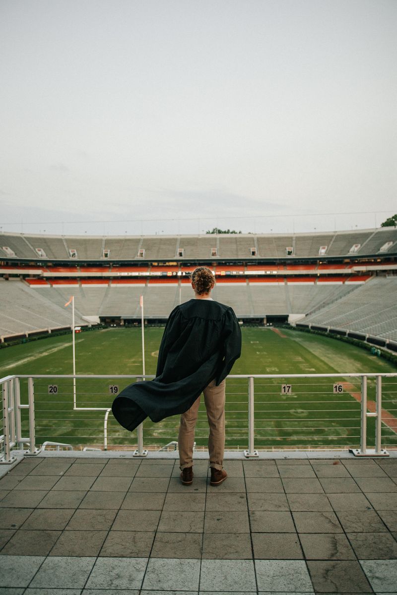 The graduate at the stadium rail, seen from behind, his gown caught in the wind