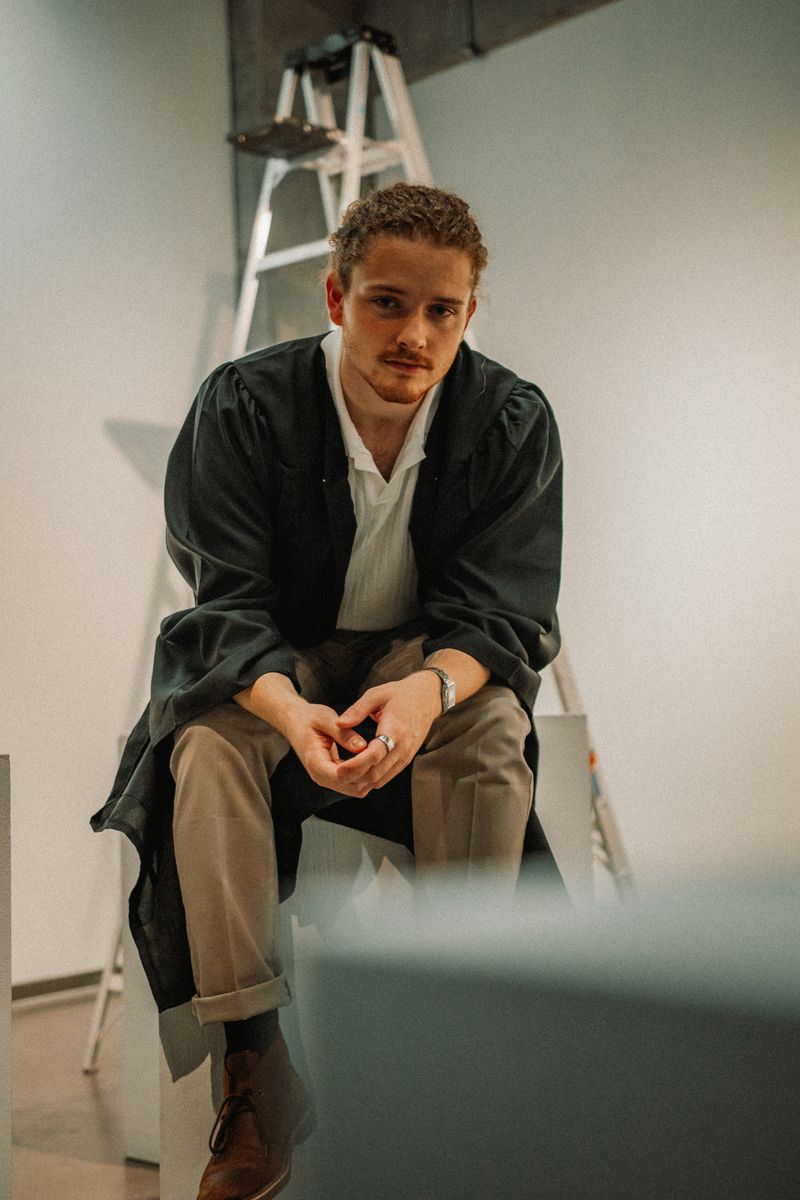 The graduate seated on a stool in front of a white wall and a ladder