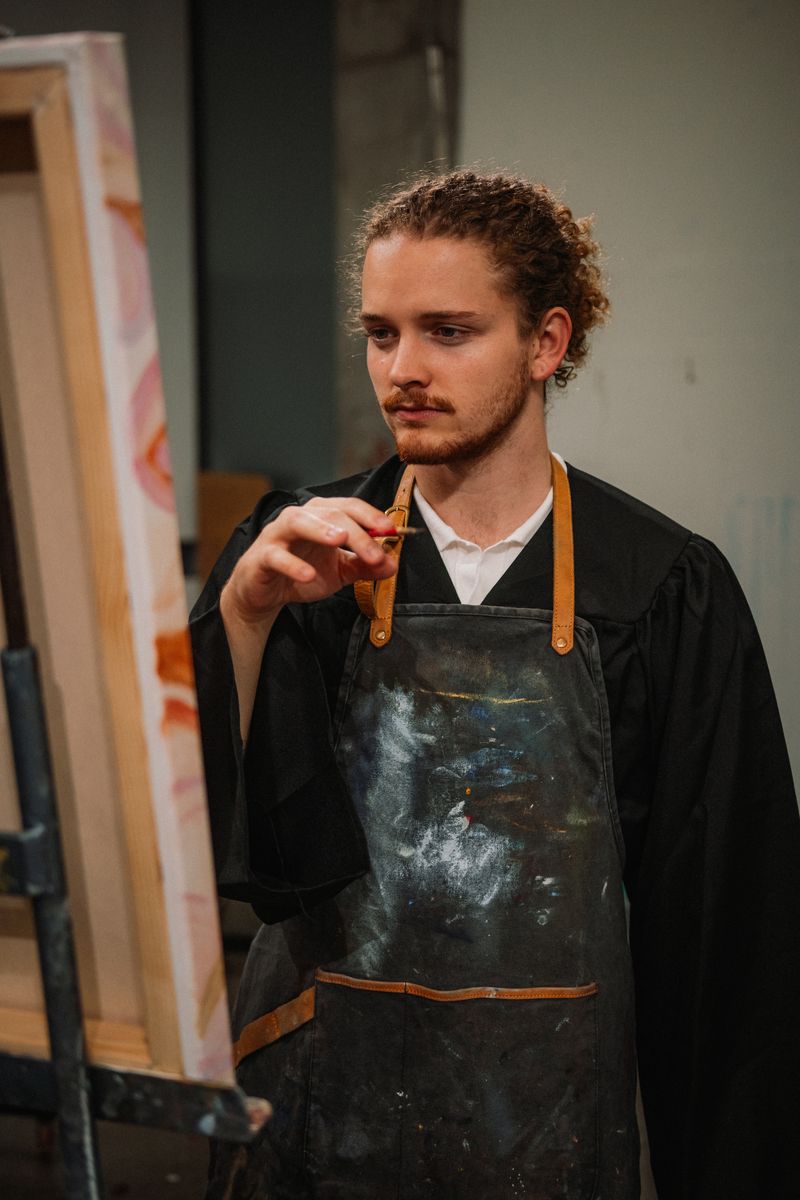 The graduate in a paint-covered apron holding a brush beside a canvas