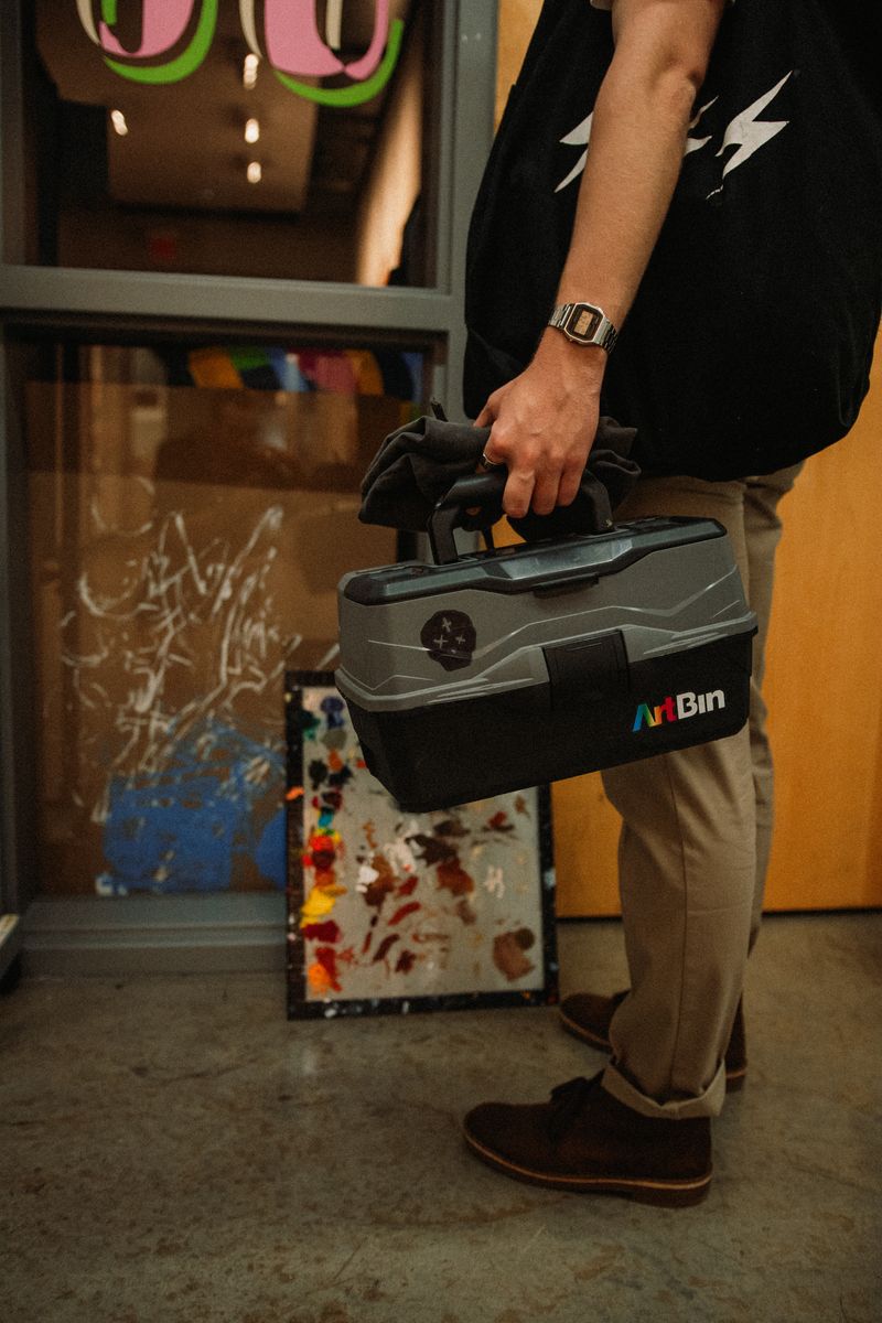 A man in sneakers carrying a paint-spattered toolbox past a mural