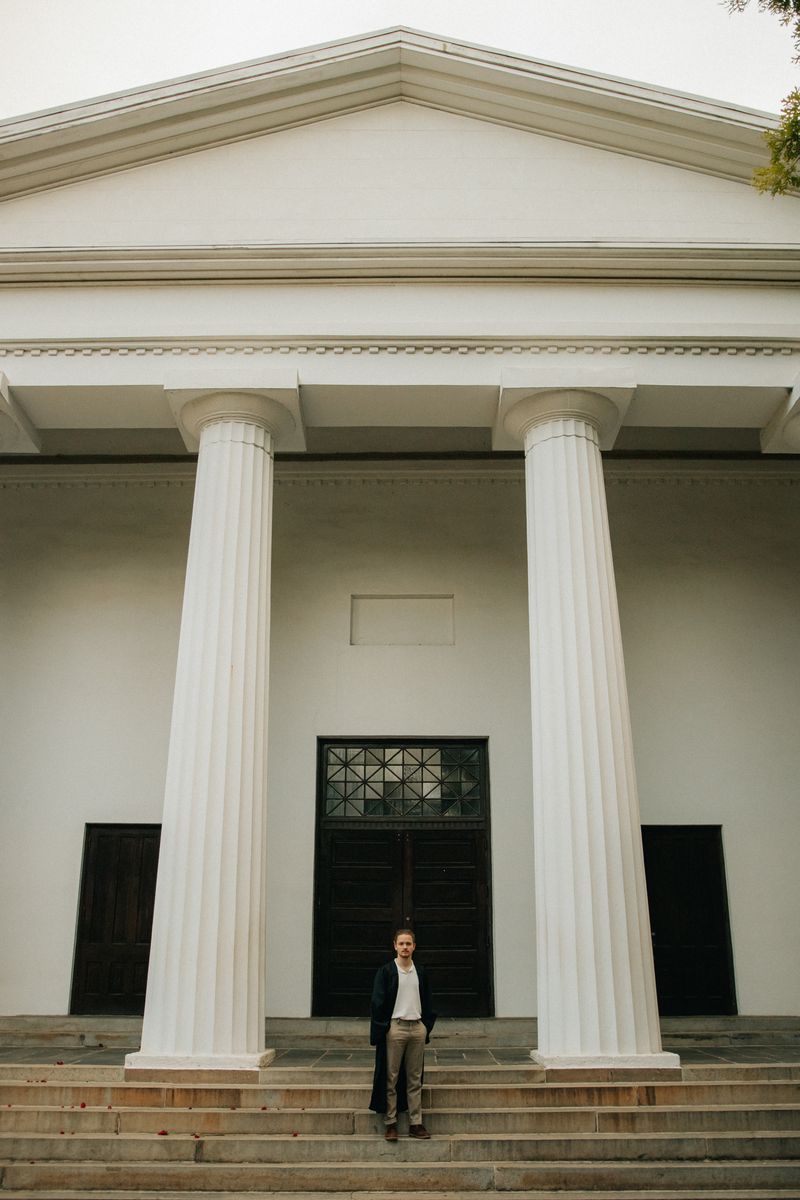 A graduate standing small on the steps of a white-columned chapel