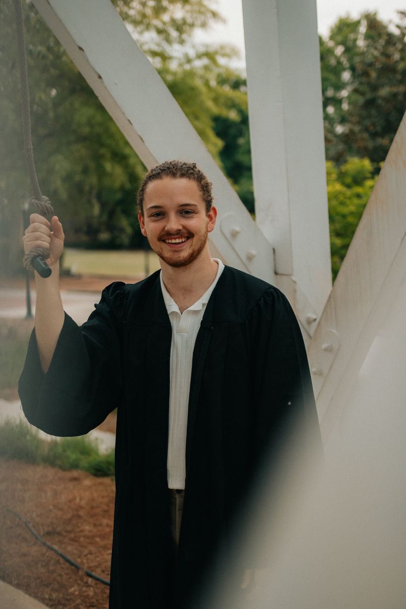 A graduate in a gown smiling under a white steel frame