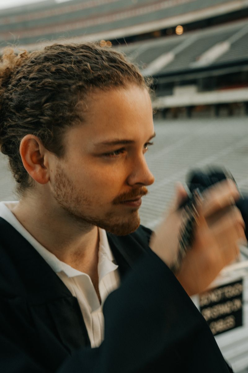 A close-up of the graduate checking a camera with the stadium behind him