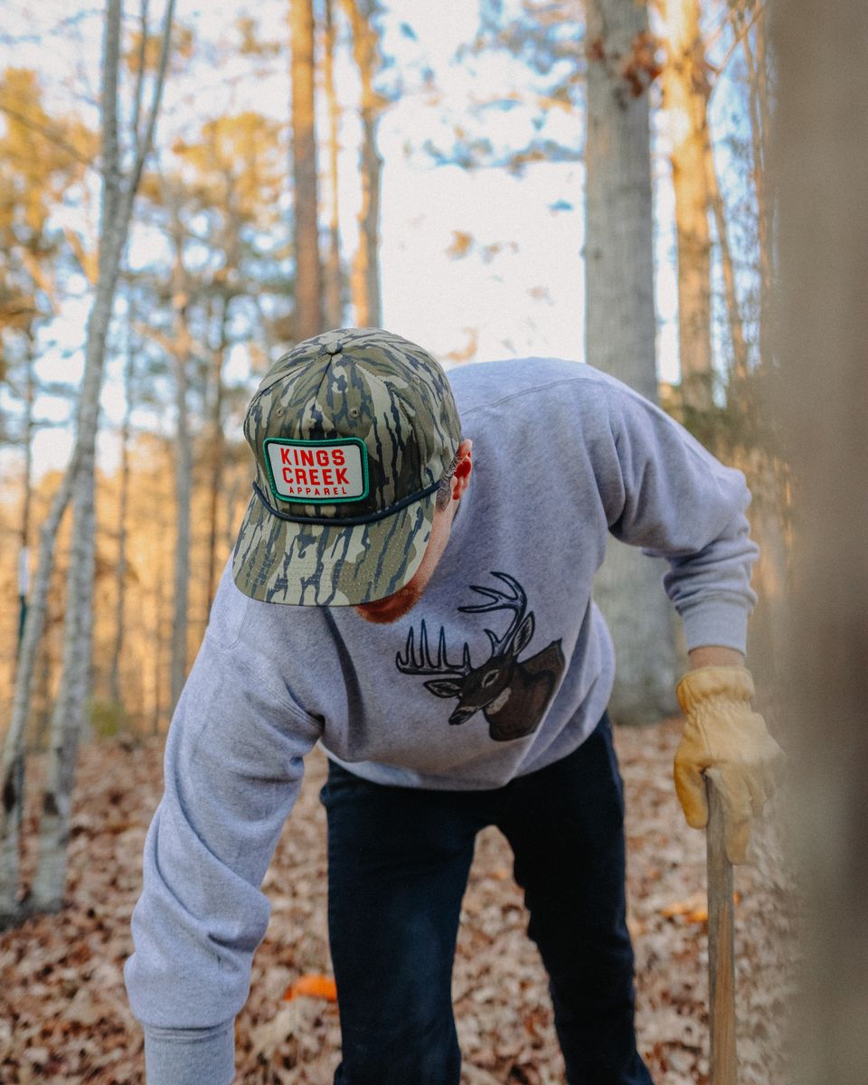 A man in a camo hat and deer sweatshirt bending to split wood