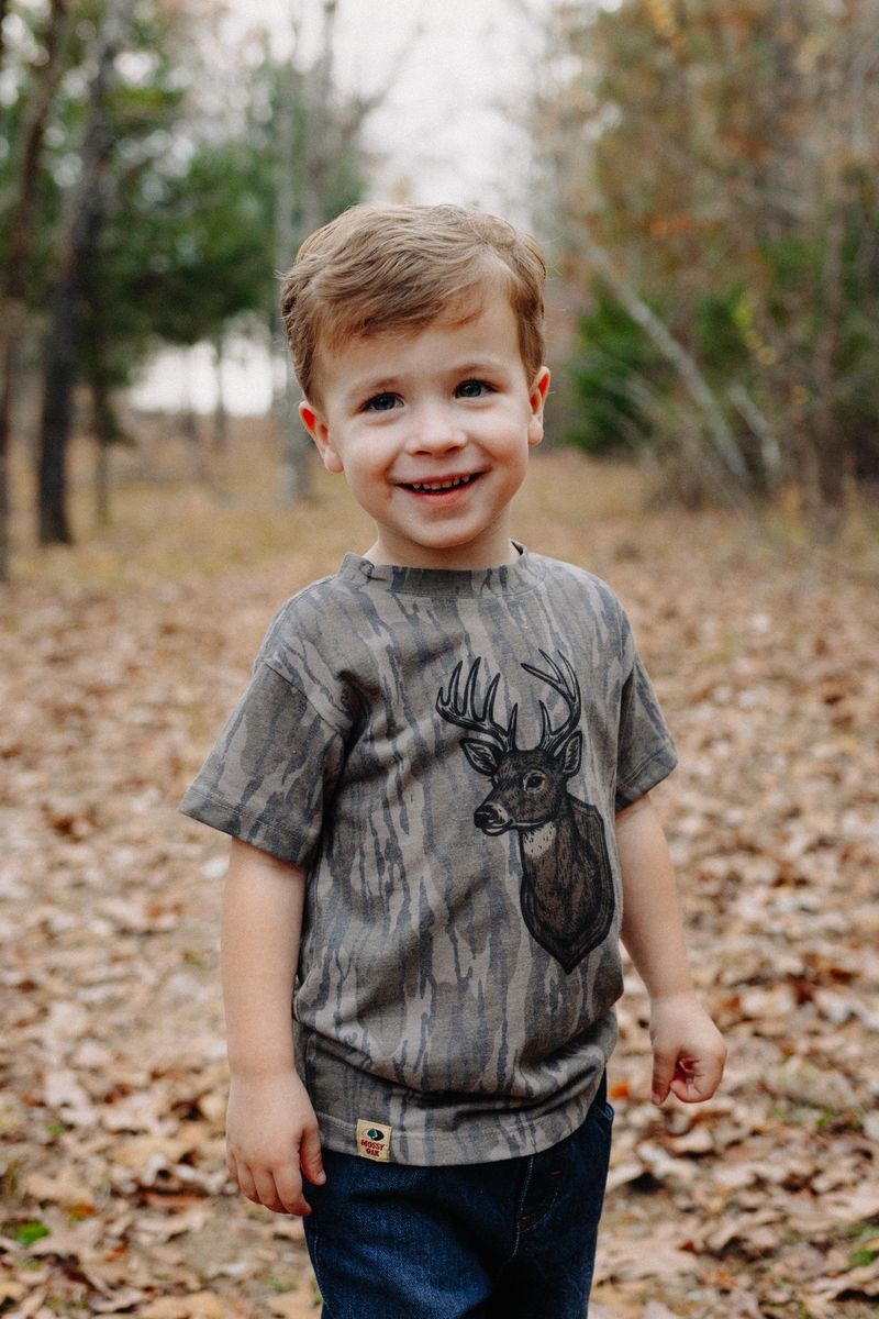 A smiling little boy in a camo deer tee standing in fallen leaves