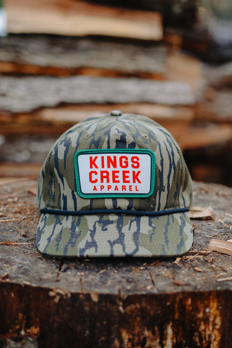 A camo rope hat with a red and white patch resting on a tree stump
