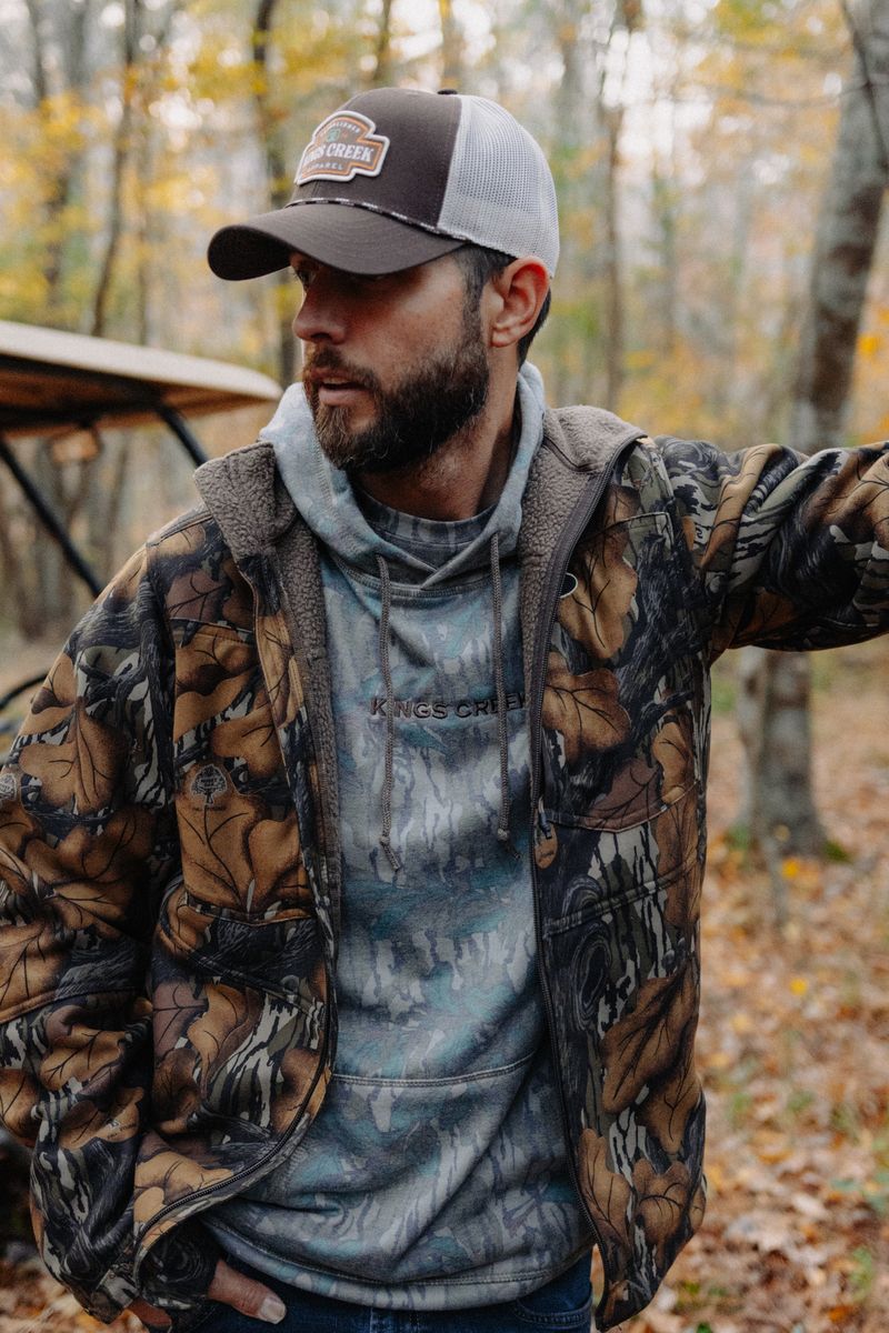 A bearded man in a camo jacket and hoodie looking off to the side