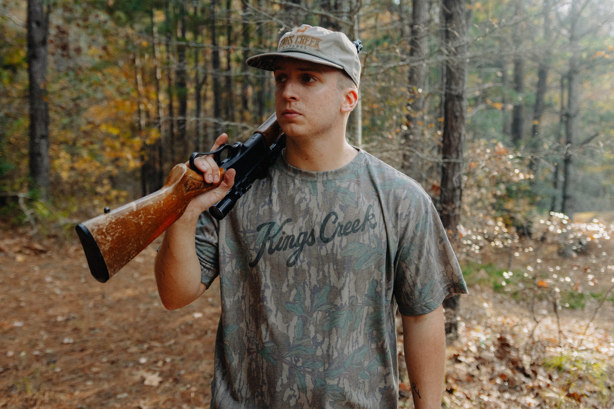 A young man in a camo tee walking through the autumn woods with a shotgun over his shoulder