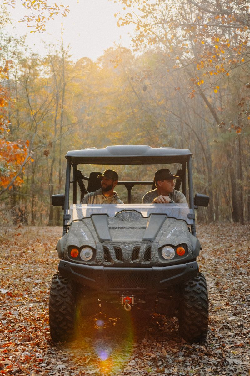Two men in a side-by-side on a leaf-covered trail with the sun behind them